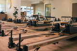 A row of Pilates reformer machines arranged in a bright, modern renew studio with wood floors and mirrored walls.