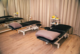 Renew treatment room with two black padded massage tables, mauve pillows, a small white storage cart, and beige curtains in the background.