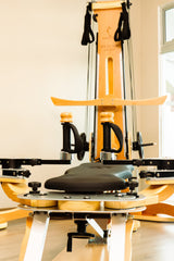 A wooden Pilates reformer machine with black straps, handles, and a padded seat, set in renew studio with light wood floors and walls.