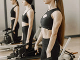 A woman in a black sports bra and black leggings stands in a gym using a Pilates reformer machine, with her reflection visible in a mirror behind her.