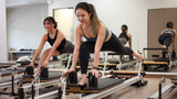 Two women performing a pilates exercise on reformer machines in a bright fitness studio.