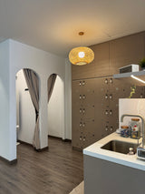 Renew modern kitchen interior with a round woven pendant light hanging above a white countertop and sink. The cabinetry is a warm taupe-brown, and the room includes two arched doorway openings with gray curtains and wood-look flooring.