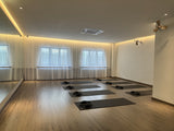 Renew studio with light wood floors, white walls, large windows with sheer curtains, and several black yoga mats arranged in rows on the floor.