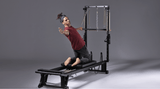 A man in a red shirt and dark pants is kneeling on a black Pilates reformer machine, holding a resistance band. The scene is shot in a studio against a solid gray background.