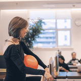 A woman in a black long-sleeve top holds an orange exercise balloon while working at a desk in a bright office or gym space.