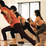 A group of people in a yoga or fitness studio performing a lunge pose, with one person in the foreground wearing a coral-pink t-shirt and dark leggings, another in a black top and leggings, and others in the background near large windows.